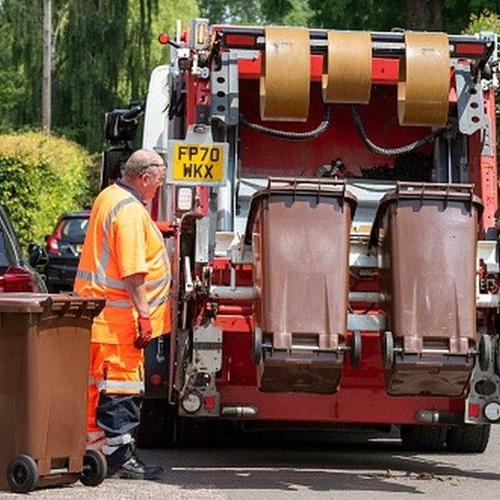 Garbage truck unloading bins with a worker in an orange safety vest.