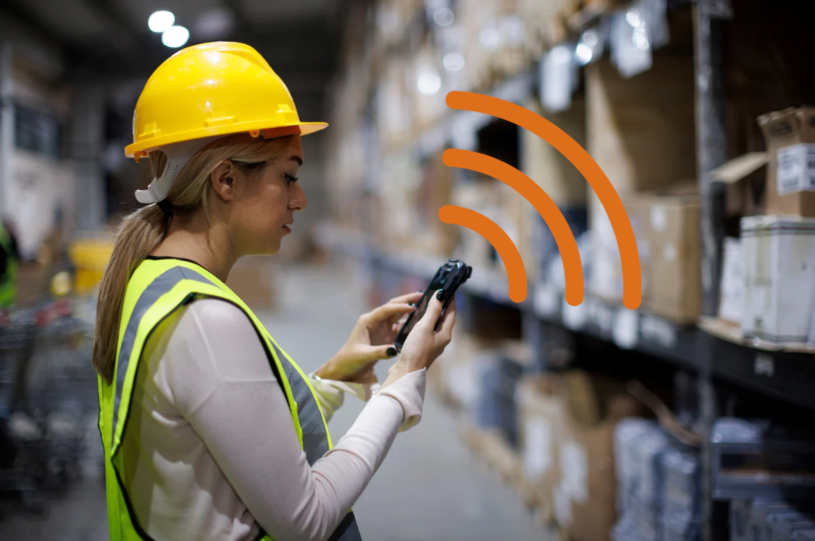 Person in a warehouse wearing a yellow hard hat and safety vest, holding a device with orange signal icons.